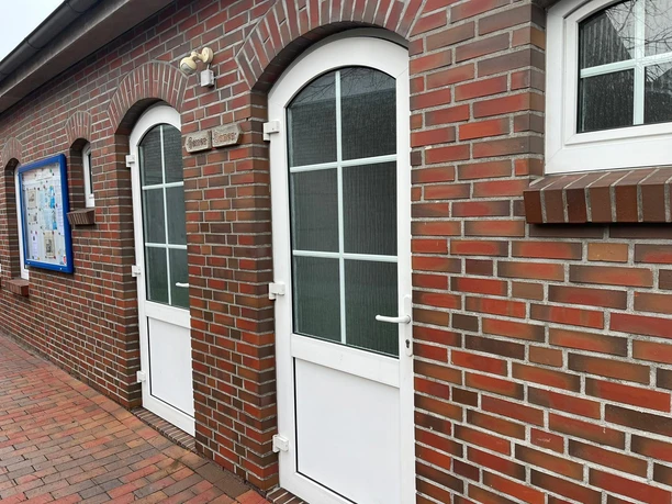 Öffentliche Toilette, Damen, Herren.jpeg Das Bild zeigt zwei nebeneinanderliegende Eingangstüren aus weißem Kunststoff in einer Backsteinfassade. Die Türen sind mit einem Schild mit der Aufschrift "Herren" und "Damen" versehen. Links davon befindet sich eine Informationstafel.