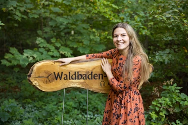 Willkommen auf dem Waldbaden-Pfad Frau in orangefarbenem Kleid steht lächelnd neben einem hölzernen Schild mit der Aufschrift "Waldbaden".