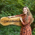 Willkommen auf dem Waldbaden-Pfad Frau in orangefarbenem Kleid steht lächelnd neben einem hölzernen Schild mit der Aufschrift "Waldbaden".