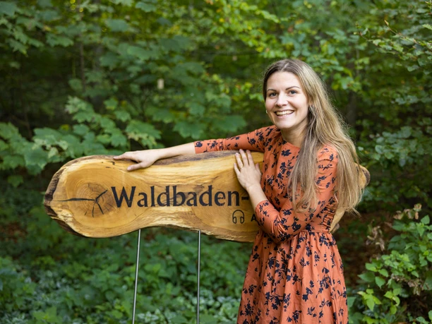 Willkommen auf dem Waldbaden-Pfad Frau in orangefarbenem Kleid steht lächelnd neben einem hölzernen Schild mit der Aufschrift "Waldbaden".