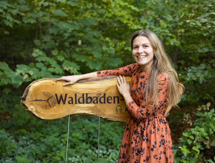 Willkommen auf dem Waldbaden-Pfad Frau in orangefarbenem Kleid steht lächelnd neben einem hölzernen Schild mit der Aufschrift "Waldbaden".