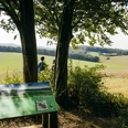 Panoramatafel Weg der Blicke Panoramatafel von "Weg der Blicke" unter Bäumen, mit Blick auf weite Felder und zwei Personen.