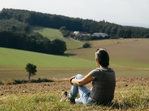Aussicht Lange Wand Person sitzt entspannt auf einer Wiese und blickt in hügelige Landschaft mit Wäldern und Feldern.