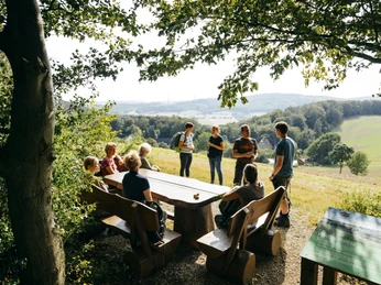 Sitzgruppe Familienplatz Gruppe von Menschen sitzt an einer großen Holztischgruppe im Grünen mit Blick auf die Landschaft.