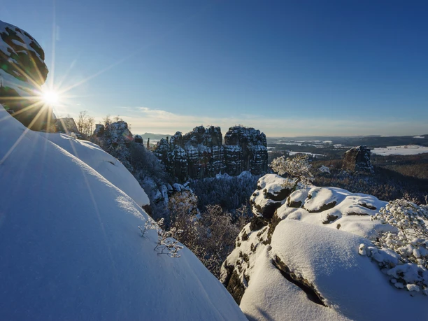 Besneeuwde Schrammsteine bij zonsopgang, de zon schijnt over besneeuwde heuvels, helderblauwe lucht op de achtergrond.