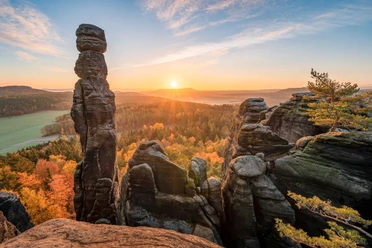Rotsformaties van de Pfaffenstein bij zonsopgang, omgeven door herfstkleurige bossen, met een heldere hemel en zachte zonnestralen.