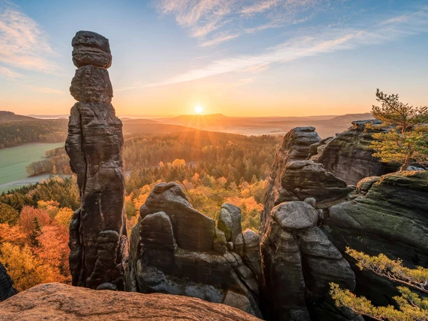 Sonnenaufgang am Pfaffenstein Felsformationen des Pfaffenstein im Sonnenaufgang, umgeben von herbstlich gefärbtem Wald, mit einem klaren Himmel und sanften Sonnenstrahlen.