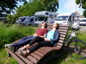 Paar auf Holzliege entspannt nahe Wohnmobilen unter blauem Himmel. Fahrrad mit Helm daneben.