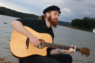 Lennon-Presse-Web.jpg Mann mit Gitarre und Mütze an einem Steg eines Sees.