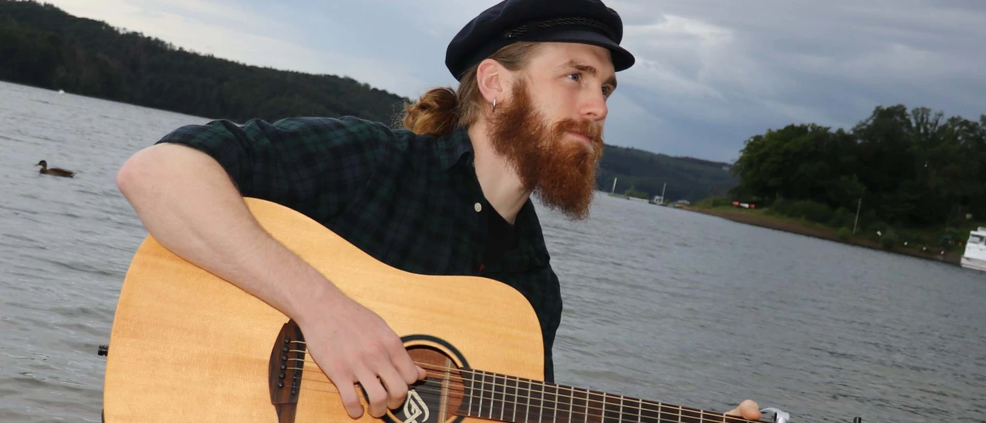 Lennon-Presse-Web.jpg Mann mit Gitarre und Mütze an einem Steg eines Sees.