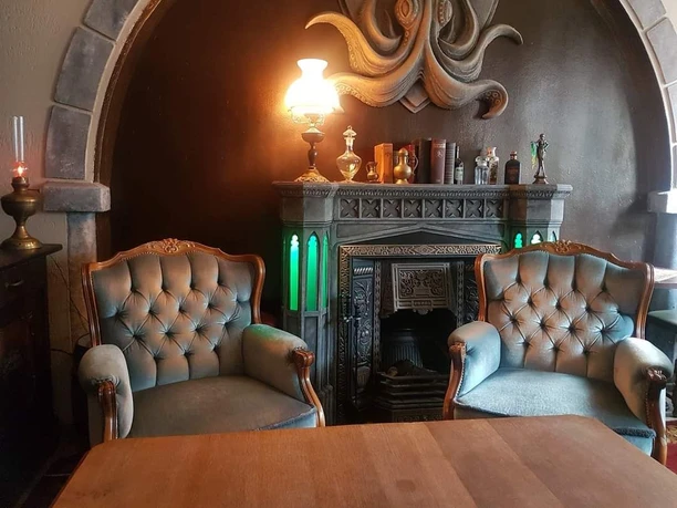 Grotesque Absinth-Bar Zwei große gepolsterte Sessel stehen an einem Tisch. An der Wand dahinter befindet sich ein Kaminofen, auf dem mehrere alte Bücher, Gefäße und eine brennende Lampe mit Lampenschirm stehen.