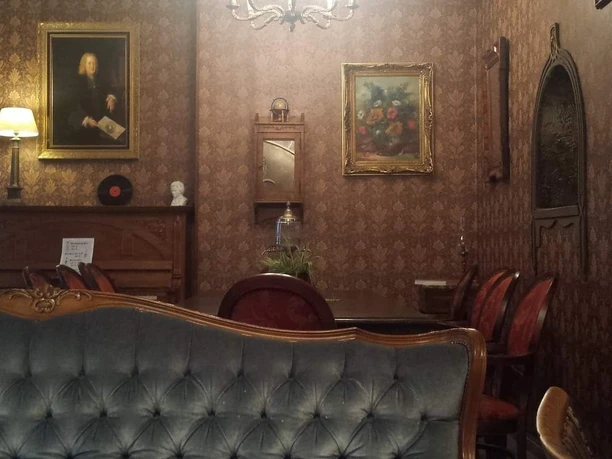 An einer tapezierten Wand hängen zwei Gemälde mit goldenem Rahmen. Unter einem davon steht ein altes Klavier aus Holz. Ein Kronleuchter hängt von der Decke. Im Vordergrund steht ein gepolstertes Sofa.