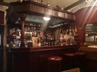 Grotesque Absinth-Bar Kleine Theke aus dunklem Holz, in der sich Regale mit Gläsern und verschiedenen Flaschen befinden. Vor der Theke stehen mehrere Barhocker.