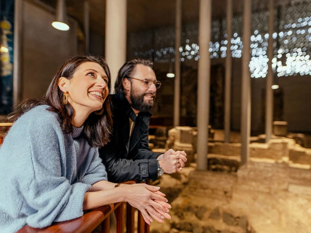 Kolumba Two smiling people admire the impressive architecture of historic stone walls from a warmly lit room - a moment of shared discovery and amazement.