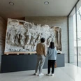 Kolumba Im Kolumba Museum in Köln betrachten zwei Personen ein großes, antikes Relief an einer modernen Ausstellungswand.In the Kolumba Museum in Cologne, two people look at a large, antique relief on a modern exhibition wall.