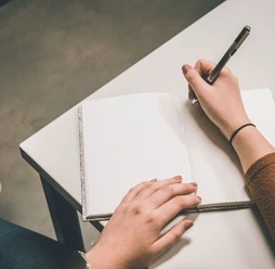 Kreatives Schreiben Eine Hand hält einen Stift und fängt an in ein leeres Buch zu schreiben, das auf einem Tisch abgelegt ist.