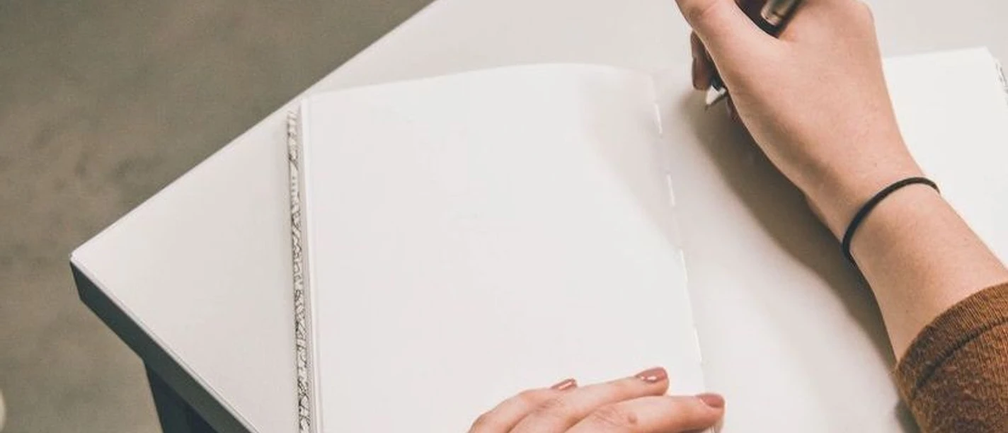 Kreatives Schreiben Eine Hand hält einen Stift und fängt an in ein leeres Buch zu schreiben, das auf einem Tisch abgelegt ist.