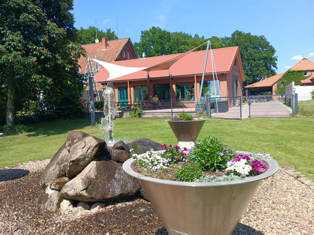 Auszeithof Otersen Ein ländliches Anwesen mit roten Backsteingebäuden, Gartenanlage und einem zentralen Springbrunnen.