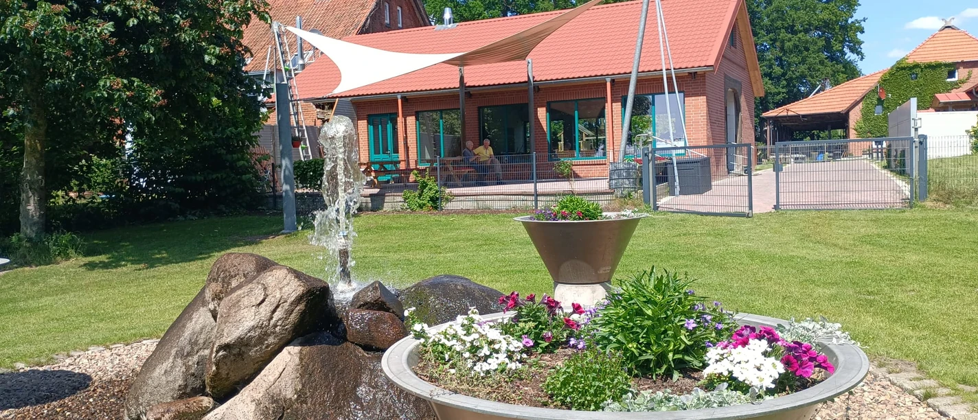 Auszeithof Otersen Ein ländliches Anwesen mit roten Backsteingebäuden, Gartenanlage und einem zentralen Springbrunnen.