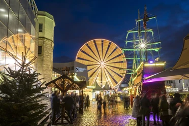 Cologne harbor Christmas market Christmas market at night with a large, brightly lit Ferris wheel in the background and a detailed, festively decorated pirate ship in the foreground. People stroll along the illuminated cobblestone path, surrounded by festively decorated stalls.