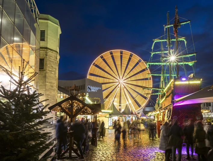 Cologne harbor Christmas market Weihnachtsmarkt bei Nacht mit einem großen, hell beleuchteten Riesenrad im Hintergrund und einem detailreichen, festlich geschmückten Piratenschiff im Vordergrund. Menschen schlendern über den beleuchteten Kopfsteinpflasterweg, umgeben von festlich dekorierten Verkaufsständen.Christmas market at night with a large, brightly lit Ferris wheel in the background and a detailed, festively decorated pirate ship in the foreground. People stroll along the illuminated cobblestone path, surrounded by festively decorated stalls.