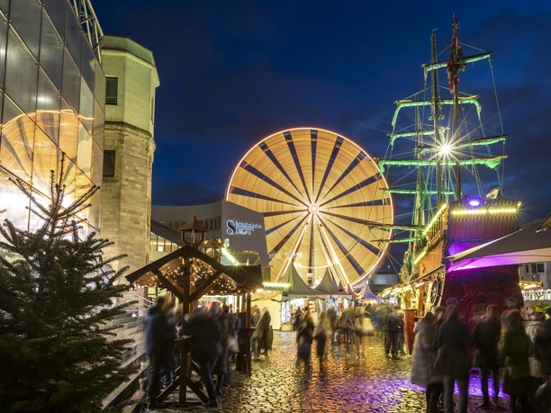 Kölner Hafen-Weihnachtsmarkt Weihnachtsmarkt bei Nacht mit einem großen, hell beleuchteten Riesenrad im Hintergrund und einem detailreichen, festlich geschmückten Piratenschiff im Vordergrund. Menschen schlendern über den beleuchteten Kopfsteinpflasterweg, umgeben von festlich dekorierten Verkaufsständen.