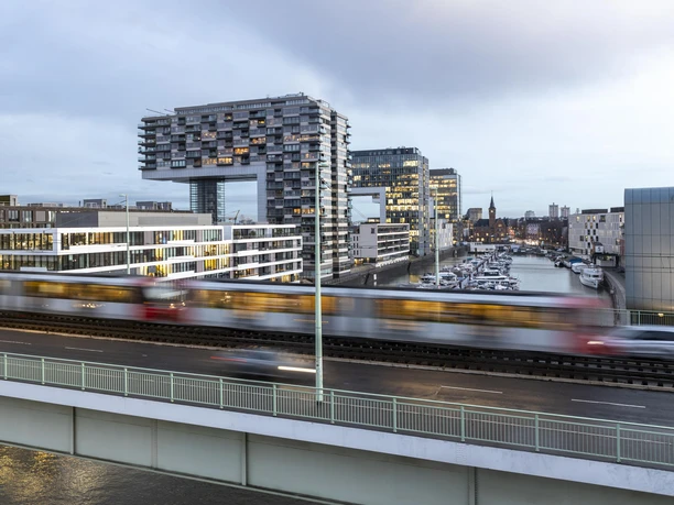 Kranhäuser Die markanten Kranhäuser im Kölner Rheinauhafen thronen modern und majestätisch entlang des Flusses. Vorne verläuft eine stark befahrene Eisenbahnbrücke, die einen lebendigen Kontrast zur stilvollen Architektur der Gebäude bildet.