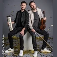 Zwei Musiker mit Keyboard und Geige, umgeben von goldenen Konfetti, sitzen entspannt auf einem Podest.