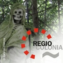 Eine steinerne Figur eines Skeletts, von einem Umhang umgeben, steht in einem von Bäumen beschatteten Bereich.