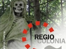 Eine steinerne Figur eines Skeletts, von einem Umhang umgeben, steht in einem von Bäumen beschatteten Bereich.