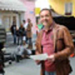 Ein Mann in Lederjacke hält eine Menükarte in einer belebten Straße mit Menschen im Hintergrund.