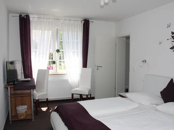 Doppelzimmer mit zwei weißen Betten, Schreibtisch, zwei Stühlen und einem Fenster mit Vorhängen.