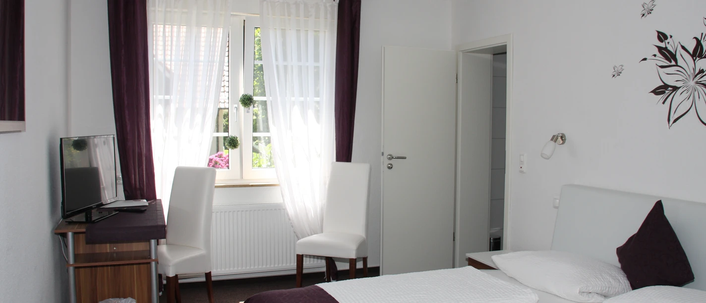 Doppelzimmer mit zwei weißen Betten, Schreibtisch, zwei Stühlen und einem Fenster mit Vorhängen.