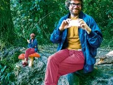 Ein lächelnder Mann sitzt in einem Wald auf einem Baumstamm, umgeben von üppigem Grün.