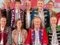 Neun Personen in traditioneller rheinischer Karnevalstracht vor buntem Hintergrund.