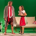 Zwei Personen in humorvoller Szene, einer in Morgenmantel und Shorts, die andere im Dirndl.