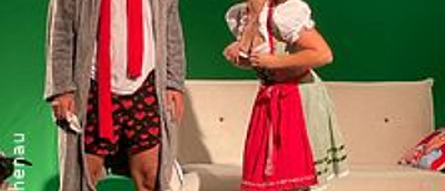 Zwei Personen in humorvoller Szene, einer in Morgenmantel und Shorts, die andere im Dirndl.
