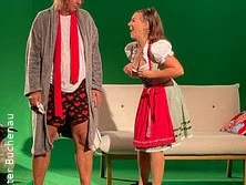 Zwei Personen in humorvoller Szene, einer in Morgenmantel und Shorts, die andere im Dirndl.