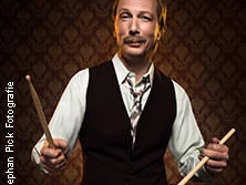 Ein Mann sitzt mit Schlagzeugstöcken in der Hand, lächelt selbstbewusst in die Kamera.