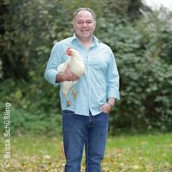 Ein Mann in hellem Hemd und Jeans steht auf grüner Wiese und hält ein weißes Huhn im Arm.