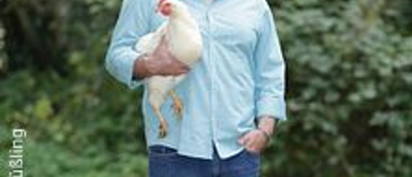 Ein Mann in hellem Hemd und Jeans steht auf grüner Wiese und hält ein weißes Huhn im Arm.
