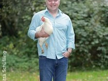 Ein Mann in hellem Hemd und Jeans steht auf grüner Wiese und hält ein weißes Huhn im Arm.