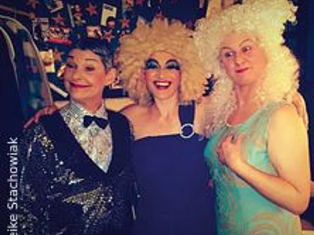 Drei Personen in farbenfroher, extravaganter Verkleidung posieren lachend vor einer Fotowand.