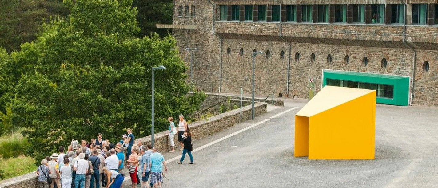 Vogelsang Führung Personengruppe steht vor modernem, gelbem Kunstwerk, historische Burg Vogelsang im Hintergrund.