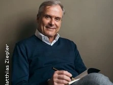 Ein älterer Mann in einem blauen Pullover sitzt lächelnd mit einem Notizbuch und Stift vor neutralem Hintergrund.