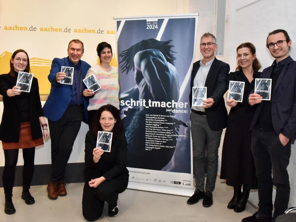 Gruppe von sieben Personen mit Plakaten des Tanzfestivals 'Schrittmacher', lächelnd vor einem Roll-Up stehend.