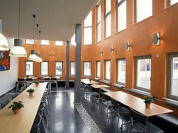 Heller, moderner Speiseraum mit hohen Decken, großen Fenstern, Tischen, Stühlen und Pflanzen.