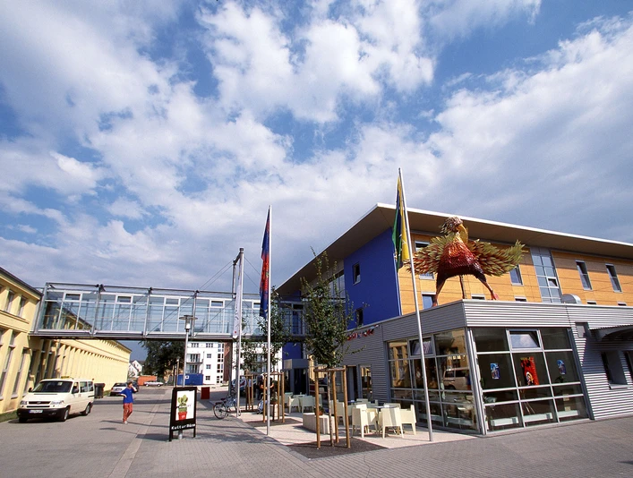 Modernes Museumsgebäude mit blauer Fassade, Glassteg und großer Skulptur davor unter blauem Himmel.