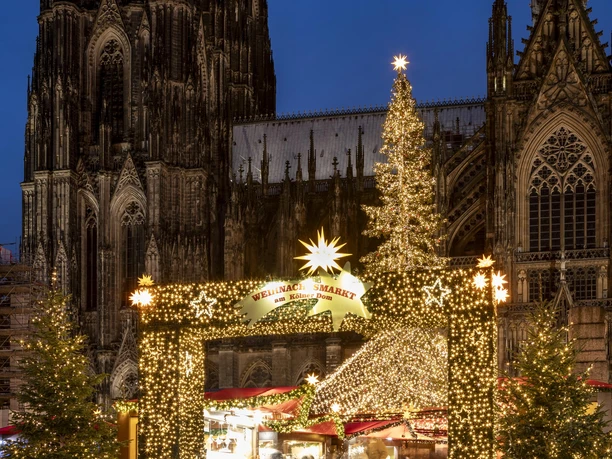 Christmas market at Cologne Cathedral The Christmas market shines in front of the majestic cathedral. Fairy lights adorn the stalls, while festive fir trees line the scene. A sparkling Christmas tree rises up in front of the impressive Gothic architecture and spreads Christmas cheer.