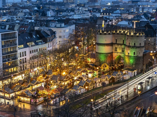 Nikolausdorf am Rudolfplatz Weihnachtsmarkt auf dem Rudolfplatz in Köln mit beleuchtetem Hahnentor in der Dämmerung.